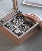 A hand opens a TAWBURY Grove 6 Slot Wooden Watch Box made from Walnut Wood, showcasing six wristwatches with various designs and bands, all neatly arranged in individual compartments beneath the glass lid.